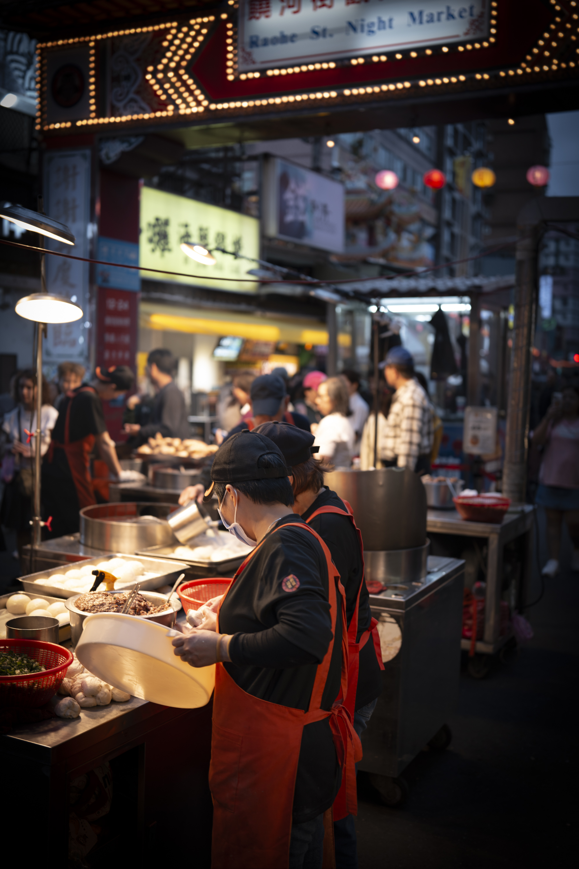 A masked vendor in a red apron prepares food at a bustling night market stall beneath glowing signage, with customers and colorful lanterns visible in the warm-lit background