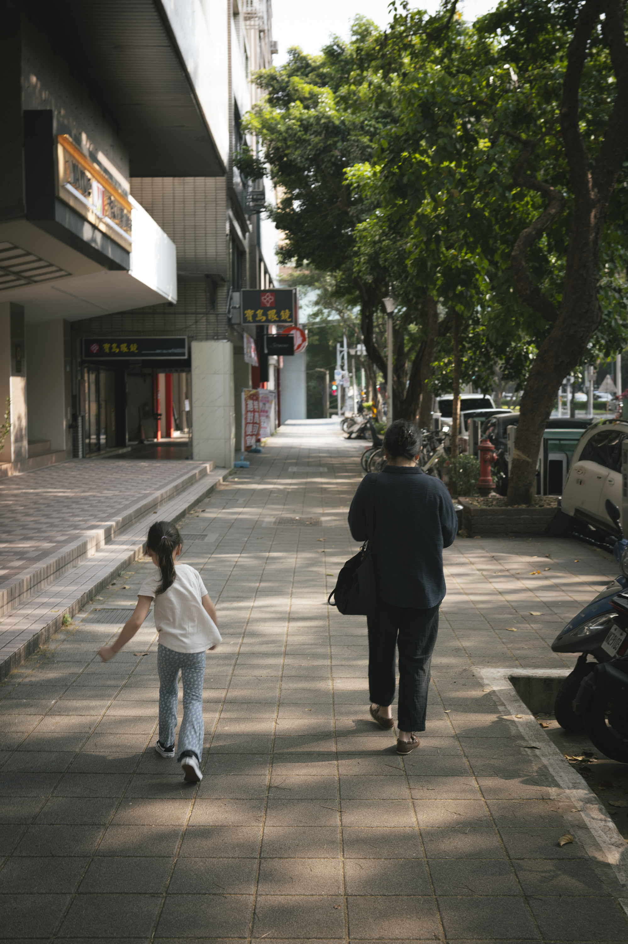 An adult and child walk down a tree-lined urban street in Taipei, passing storefronts and parked scooters under dappled sunlight filtering through leafy canopy overhead
