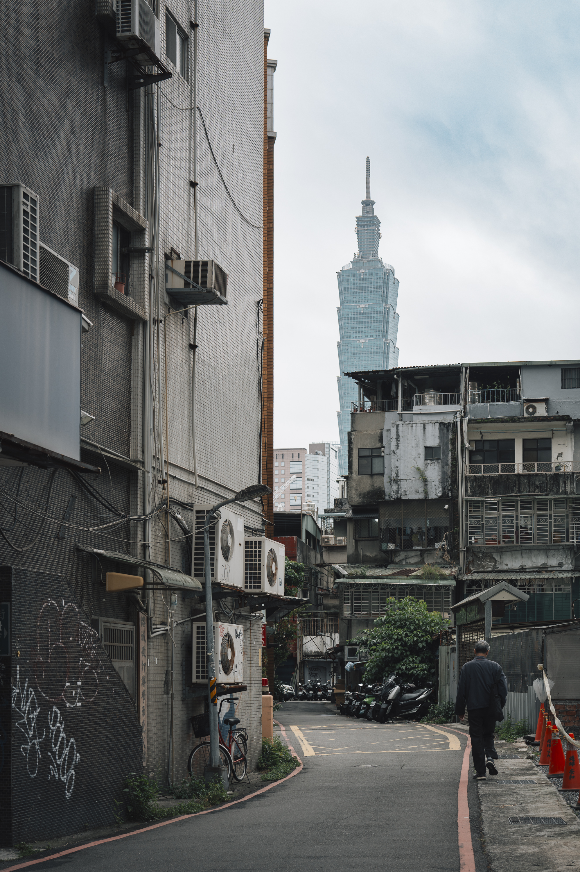 A narrow urban alley lined with weathered residential buildings frames a distant view of Taipei 101's distinctive spire rising against an overcast sky