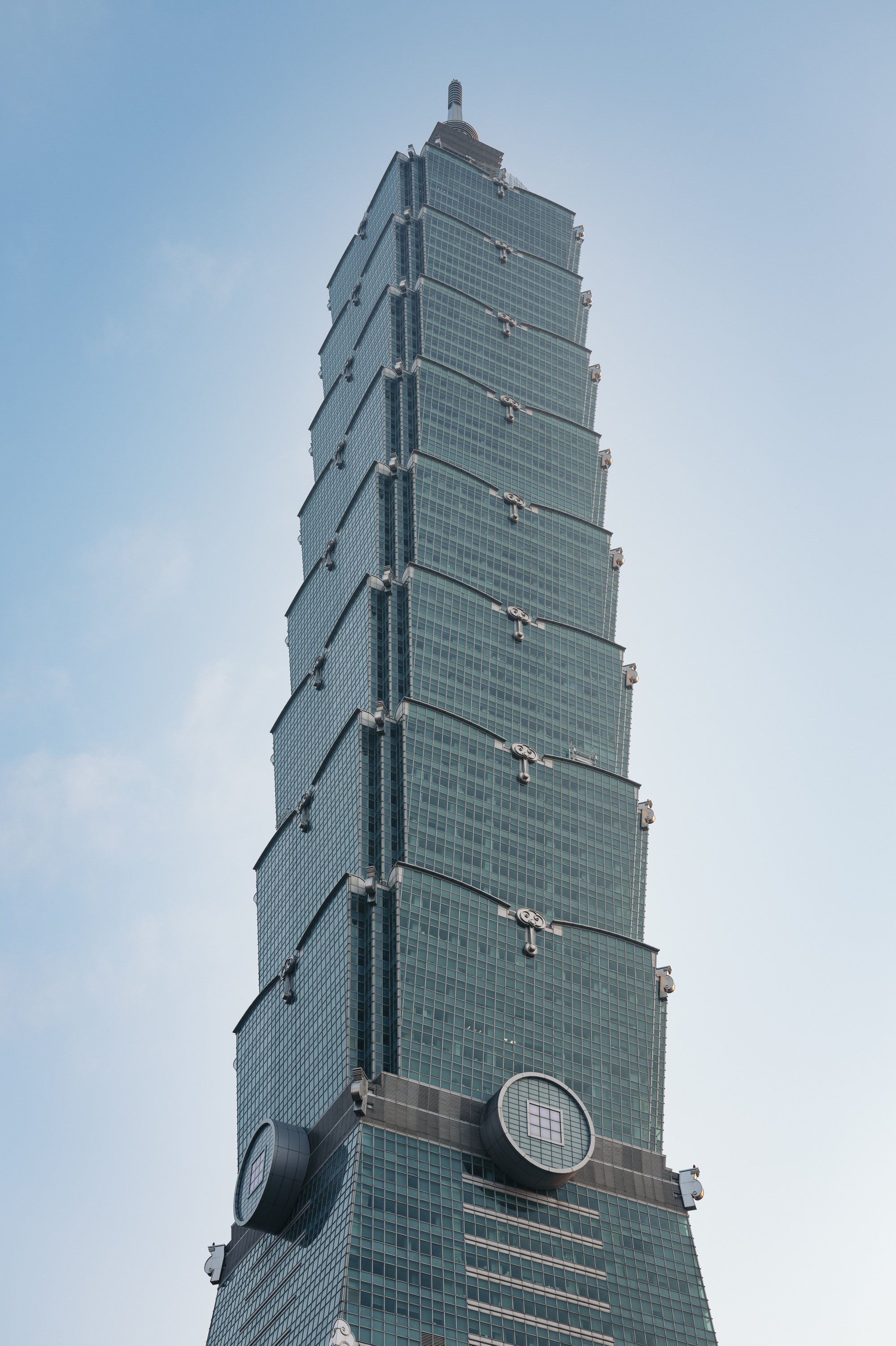 Taipei 101's upper tower section rises against a clear blue sky, its distinctive segmented green-tinted glass facade and protruding structural elements creating a tapered silhouette with visible communication spheres