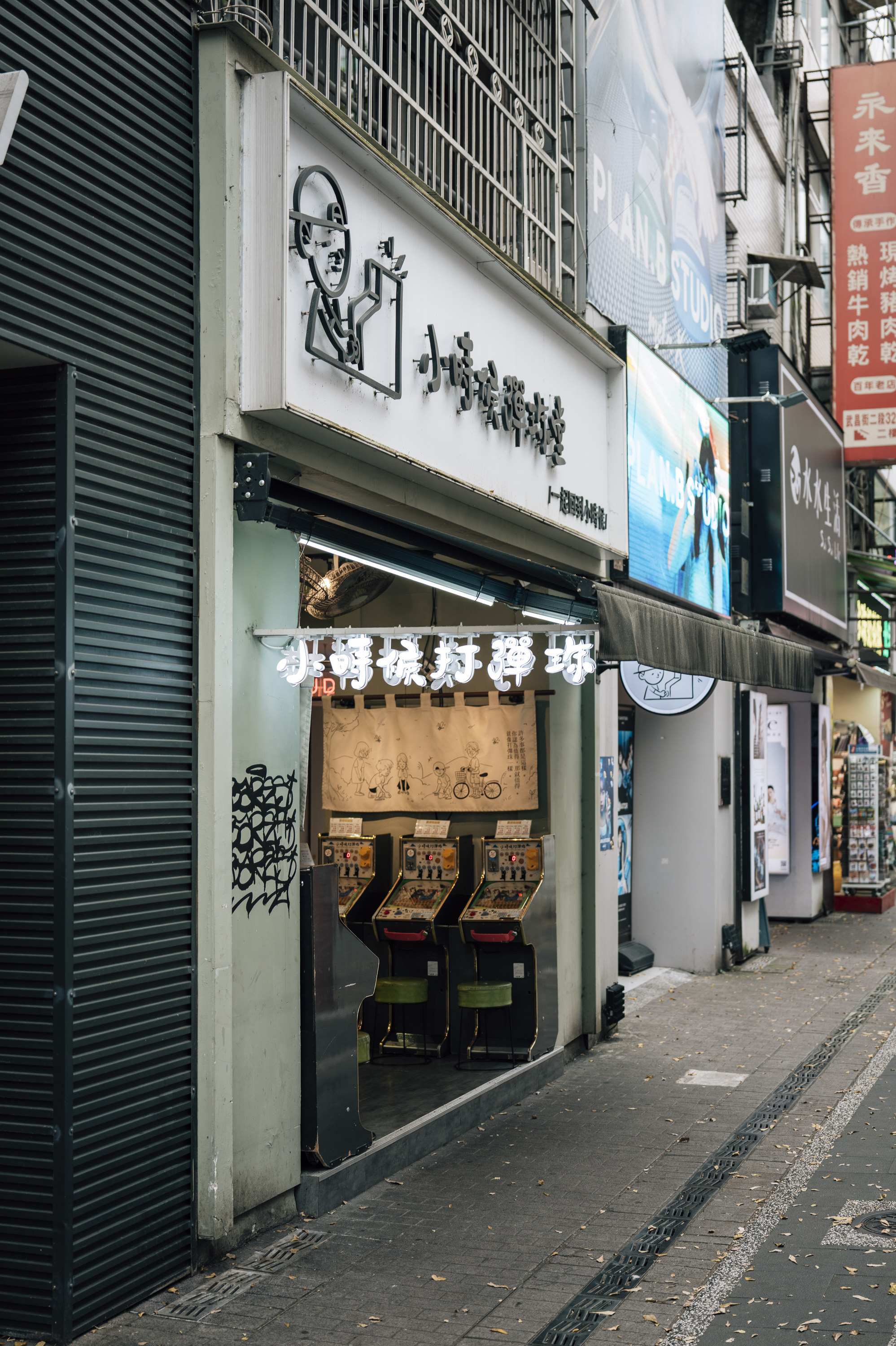 A narrow Taipei alleyway displays storefronts with Japanese signage and arcade machines visible through glass doors, surrounded by weathered metal shutters and shop signage in daylight