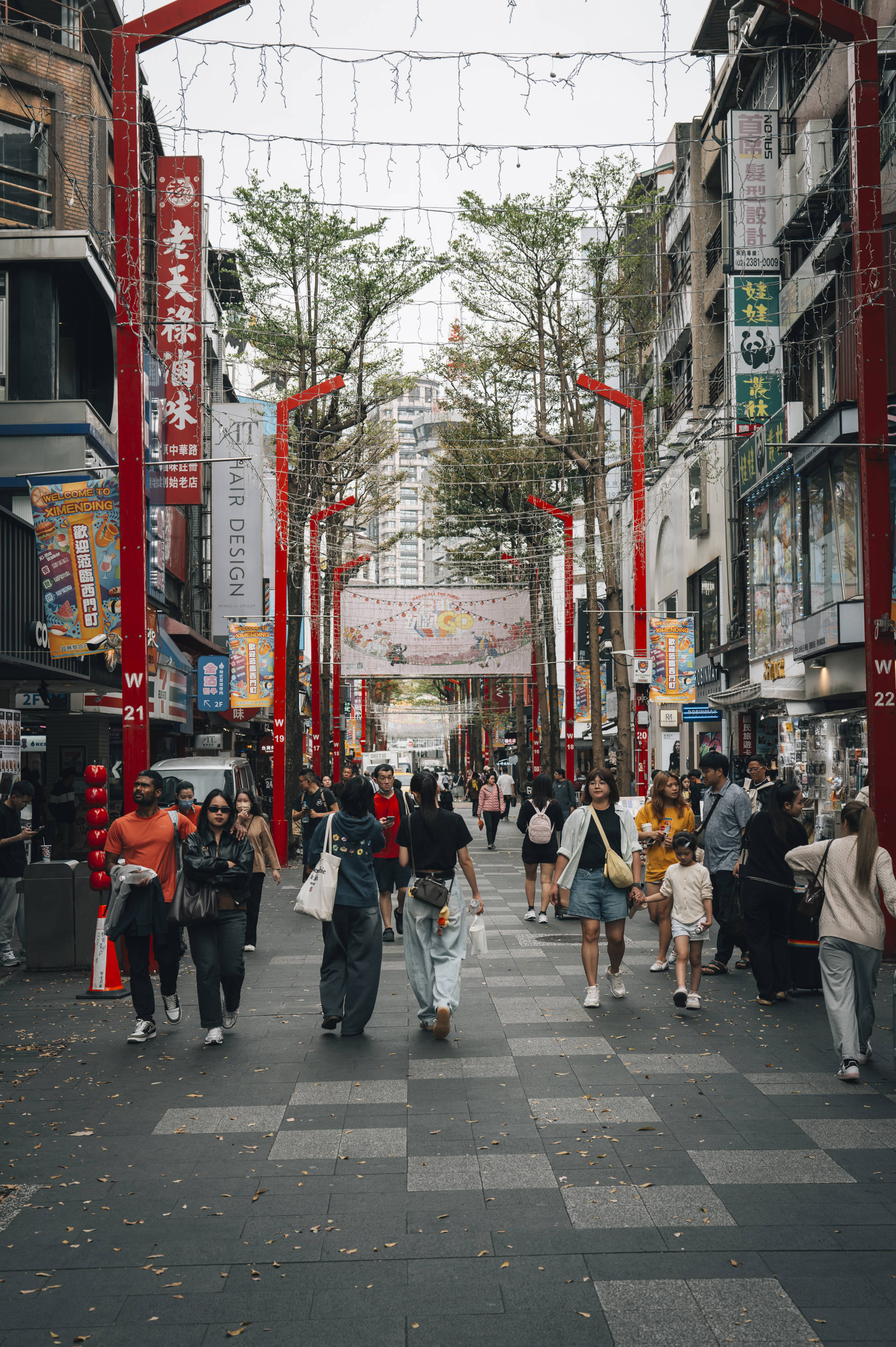 A bustling pedestrian street in Taipei lined with red decorative arches, Chinese signage, and scattered shoppers walking past commercial storefronts under overcast spring daylight.