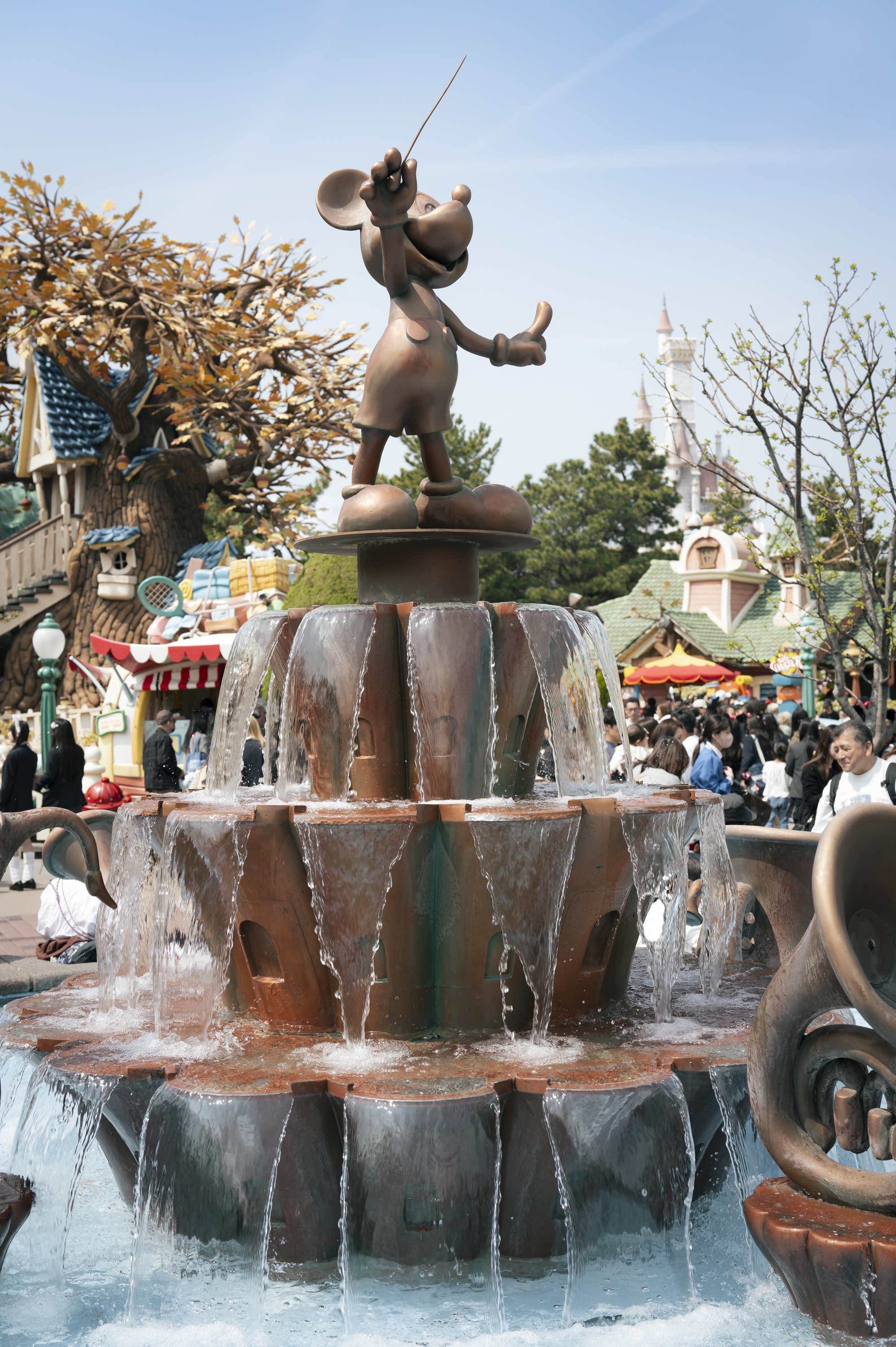 Bronze cartoon character statue stands atop a tiered fountain with cascading water in a sunny theme park plaza surrounded by visitors and attractions