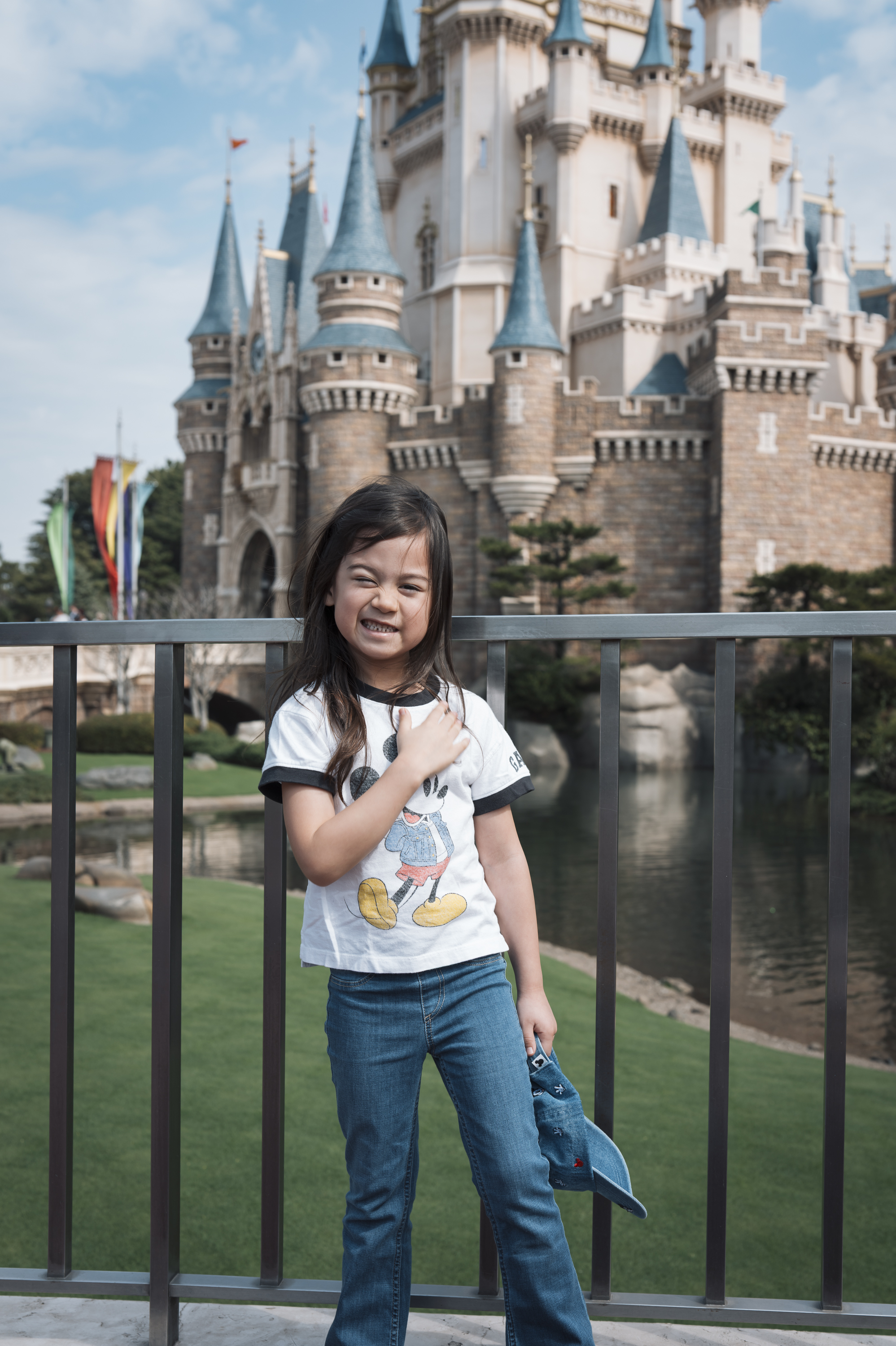 A young girl in a white graphic tee and blue jeans poses at a railing with a grand fairy tale castle framed behind her under clear skies.