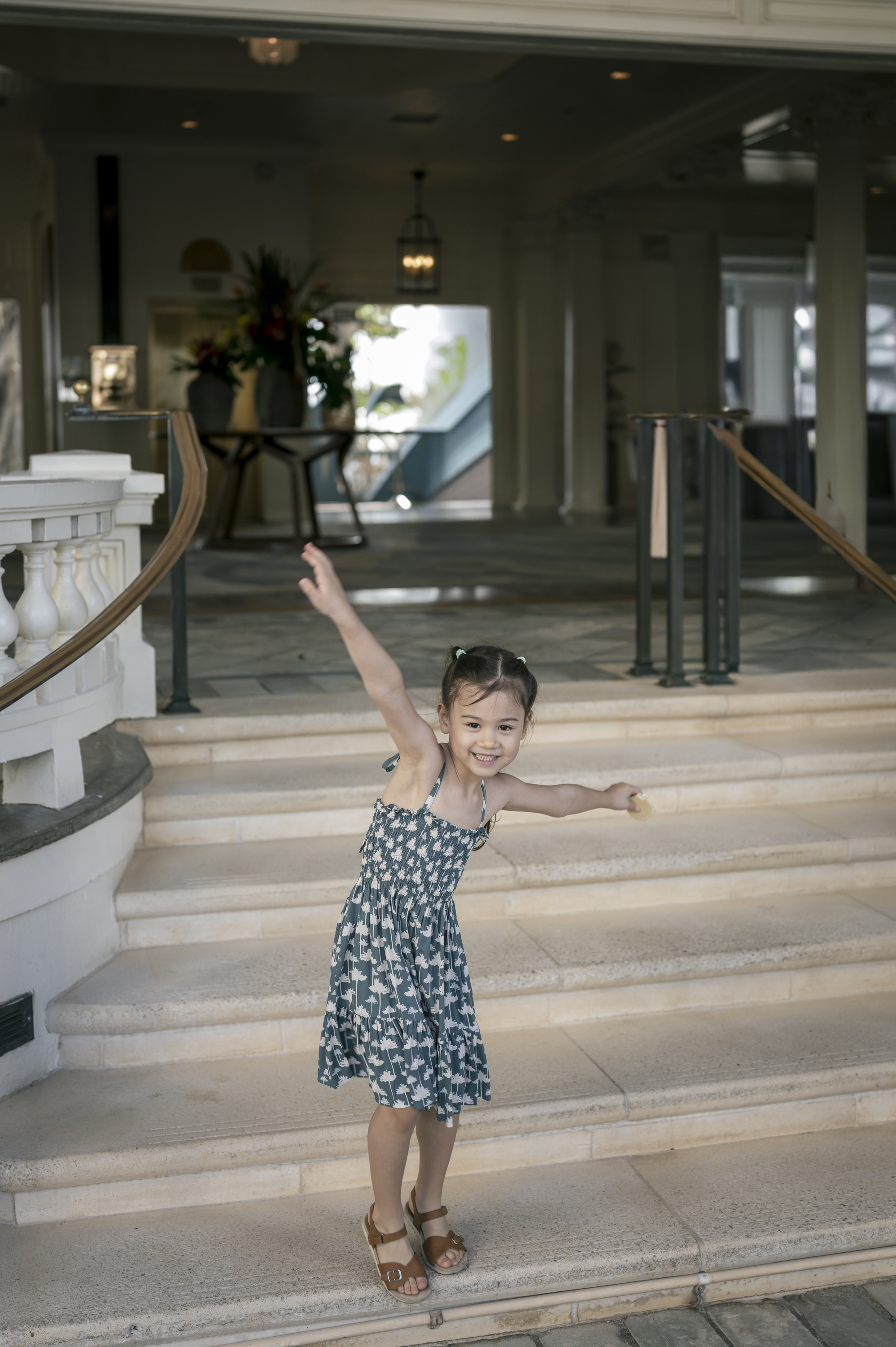 Young girl in a floral dress stands joyfully with arms raised on a wide hallway staircase inside a luxury resort, bathed in natural daylight from large windows